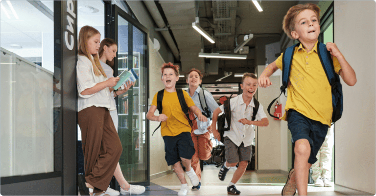 A group of children with backpacks are joyfully running down a school hallway. Two girls are standing by the wall, one reading a book and the other looking on. The hallway is brightly lit with modern decor.