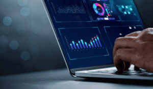 A close-up shot of a person's hands typing on a laptop keyboard. The laptop screen displays various data visualization dashboards with colorful graphs, charts, and maps against a dark blue background with glowing light effects.
