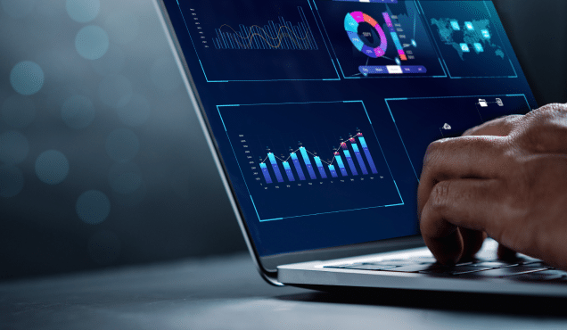 A close-up shot of a person's hands typing on a laptop keyboard. The laptop screen displays various data visualization dashboards with colorful graphs, charts, and maps against a dark blue background with glowing light effects.